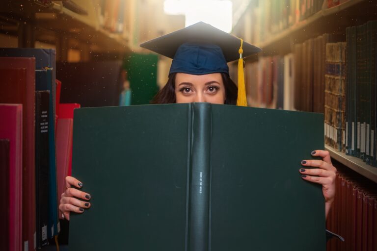 Absolventin mit Doktorhut, die ihr halbes Gesicht hinter einem Buch versteckt